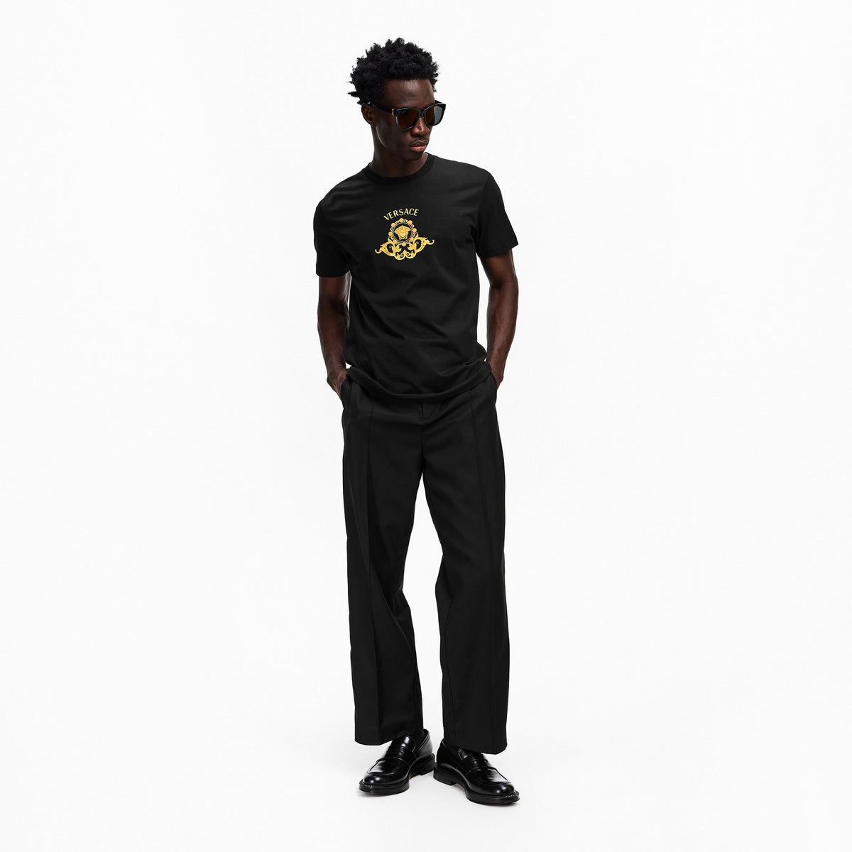 Black T-shirt with gold Medusa crest logo, worn with black trousers